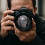 Photographie en gros plan d’un détective privé tenant un appareil photo, avec son visage dissimulé derrière l’objectif, dans lequel se reflètent deux hommes échangeant un paquet dans une rue.